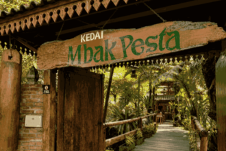 Wooden sign reading 'Kedai Mbak Pesta' over a rustic doorway leading to a leafy tropical cafe path.