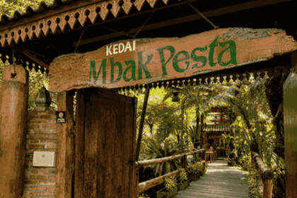 Wooden sign reading 'Kedai Mbak Pesta' over a rustic doorway leading to a leafy tropical cafe path.