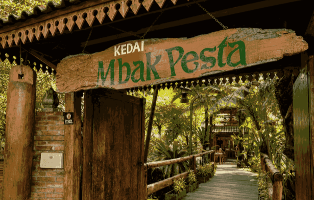Wooden sign reading 'Kedai Mbak Pesta' over a rustic doorway leading to a leafy tropical cafe path.