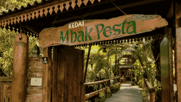 Wooden sign reading 'Kedai Mbak Pesta' over a rustic doorway leading to a leafy tropical cafe path.