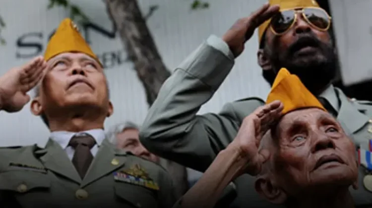 Three military personnel in dress uniforms salute while looking upward, two wearing yellow folded hats and one with sunglasses, during a ceremony.