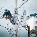 Two utility workers in blue uniforms and helmets repair electrical wires atop a tall utility pole, with a bucket truck nearby.