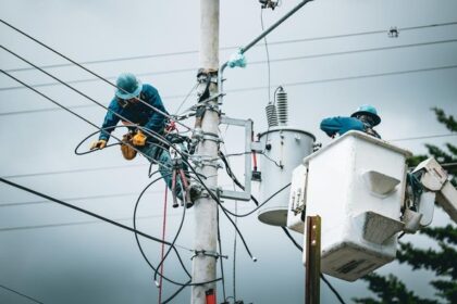 Two utility workers in blue uniforms and helmets repair electrical wires atop a tall utility pole, with a bucket truck nearby.