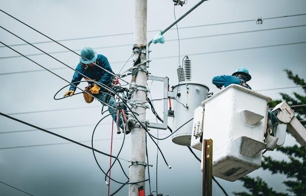 Two utility workers in blue uniforms and helmets repair electrical wires atop a tall utility pole, with a bucket truck nearby.
