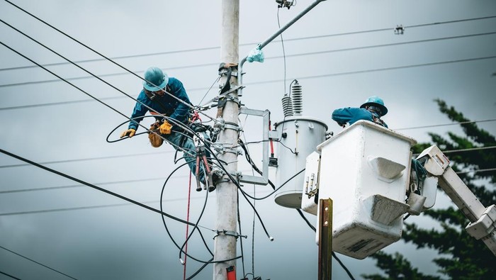 Two utility workers in blue uniforms and helmets repair electrical wires atop a tall utility pole, with a bucket truck nearby.