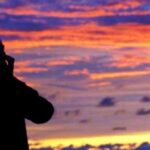 Silhouette of a man talking on a mobile phone against a vibrant sunset sky with orange and purple clouds.