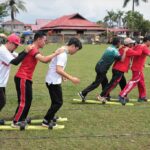 Group of seven people in red and white shirts balancing on tandem planks outdoors, linking arms for a team-building exercise.