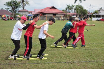 Group of seven people in red and white shirts balancing on tandem planks outdoors, linking arms for a team-building exercise.