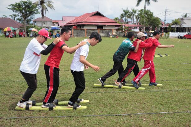 Group of seven people in red and white shirts balancing on tandem planks outdoors, linking arms for a team-building exercise.