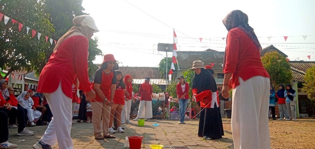 balap gelas air di atas piring - Mabur.co Group of people in red shirts playing a cup-and-string telephone game outdoors at a community event.