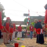 Group of people in red shirts playing a cup-and-string telephone game outdoors at a community event.