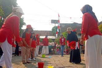 Group of people in red shirts playing a cup-and-string telephone game outdoors at a community event.
