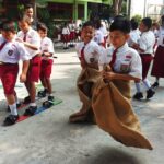 Group of elementary students in white shirts and red shorts taking part in a sack race outdoors; a girl hops in a burlap sack while classmates cheer nearby.