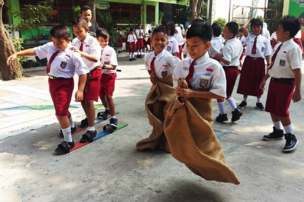 Group of elementary students in white shirts and red shorts taking part in a sack race outdoors; a girl hops in a burlap sack while classmates cheer nearby.