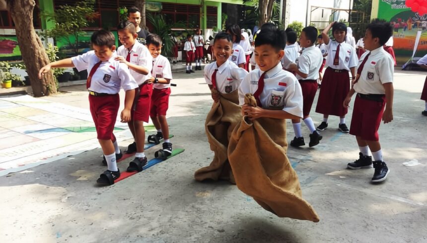 Group of elementary students in white shirts and red shorts taking part in a sack race outdoors; a girl hops in a burlap sack while classmates cheer nearby.