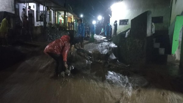 Nighttime street flooded with muddy water; people in rain gear wade through debris and mud under streetlights.