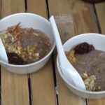 Two white bowls on a wooden table containing raw meat with pasta and vegetables, arranged for cooking or serving.
