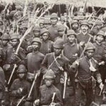 Black-and-white photo of a large group of soldiers in vintage uniforms holding rifles, posing for a military portrait in a dense formation.