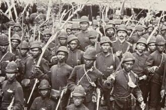 Black-and-white photo of a large group of soldiers in vintage uniforms holding rifles, posing for a military portrait in a dense formation.