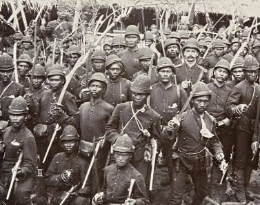 Black-and-white photo of a large group of soldiers in vintage uniforms holding rifles, posing for a military portrait in a dense formation.