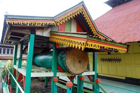bedhuk-masjid-agung-pondok-tinggi - Mabur.co Colorful traditional structure with a large ceremonial drum mounted on a green-and-red wooden platform beside a bright yellow wall and decorative eaves.
