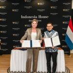 Two women stand by a table holding signed documents in front of an Indonesia House Amsterdam backdrop, with Indonesian and Dutch flags nearby.