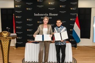Two women stand by a table holding signed documents in front of an Indonesia House Amsterdam backdrop, with Indonesian and Dutch flags nearby.