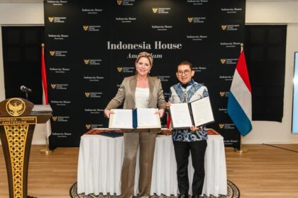 Two women stand by a table holding signed documents in front of an Indonesia House Amsterdam backdrop, with Indonesian and Dutch flags nearby.