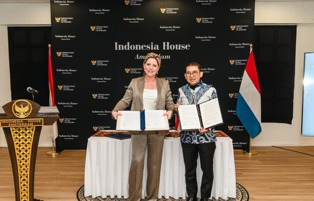 Two women stand by a table holding signed documents in front of an Indonesia House Amsterdam backdrop, with Indonesian and Dutch flags nearby.