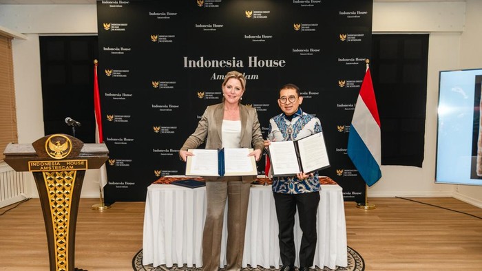 Two women stand by a table holding signed documents in front of an Indonesia House Amsterdam backdrop, with Indonesian and Dutch flags nearby.