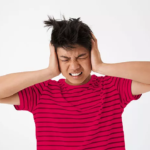 Young man in a red striped shirt covers his ears and grimaces, overwhelmed by loud noise in a bright setting.