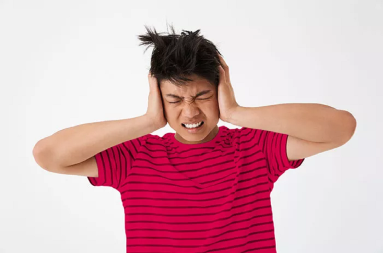 benci suara sendiri by halodoc - Mabur.co Young man in a red striped shirt covers his ears and grimaces, overwhelmed by loud noise in a bright setting.