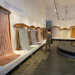 Gallery aisle with ornate carved frames holding long, colorful woven textiles; a visitor examines them.