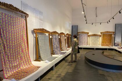 Gallery aisle with ornate carved frames holding long, colorful woven textiles; a visitor examines them.