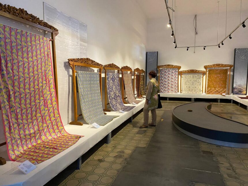 Gallery aisle with ornate carved frames holding long, colorful woven textiles; a visitor examines them.