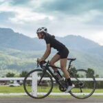 Woman cyclist wearing a white helmet and black kit riding a road bike along a rural road with mountains in the background.