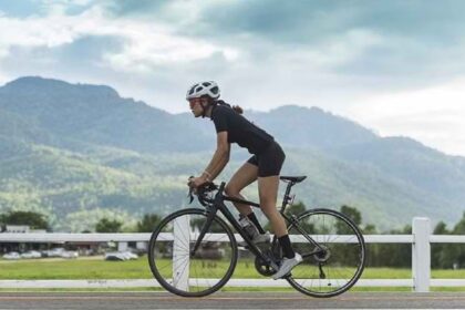 Woman cyclist wearing a white helmet and black kit riding a road bike along a rural road with mountains in the background.