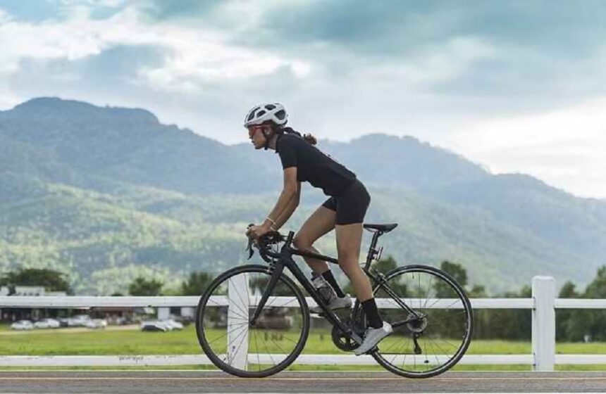 bersepeda by halodoc - Mabur.co Woman cyclist wearing a white helmet and black kit riding a road bike along a rural road with mountains in the background.