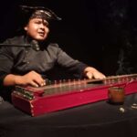 Musician in a bandana playing a red hammered dulcimer at a microphone on stage, lit against a dark background.