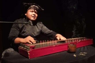 Musician in a bandana playing a red hammered dulcimer at a microphone on stage, lit against a dark background.