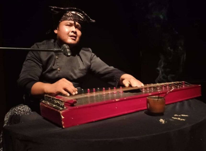Musician in a bandana playing a red hammered dulcimer at a microphone on stage, lit against a dark background.