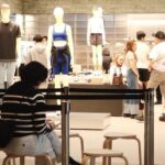 Shoppers browse a clothing store with mannequins on display and a person sitting in the foreground wearing a striped sweater.