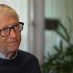 Older man with gray hair and glasses sits indoors, wearing a dark sweater over a light collared shirt, with plants in the background.