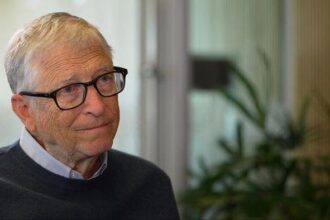 Older man with gray hair and glasses sits indoors, wearing a dark sweater over a light collared shirt, with plants in the background.