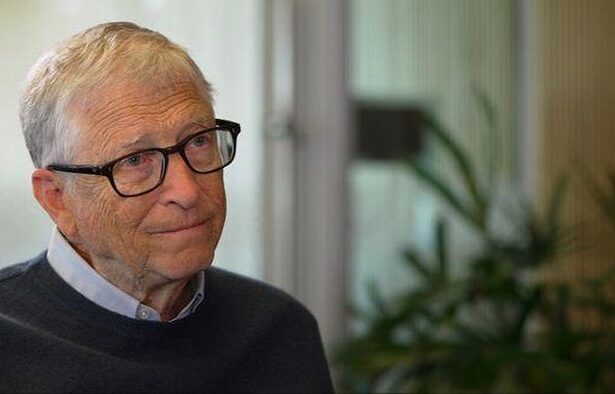 Older man with gray hair and glasses sits indoors, wearing a dark sweater over a light collared shirt, with plants in the background.