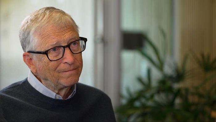 Older man with gray hair and glasses sits indoors, wearing a dark sweater over a light collared shirt, with plants in the background.