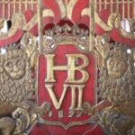 Ornate gold heraldic panel with a crown, the monogram 'HB VII', and two lion supporters on a red background.