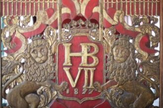 Ornate gold heraldic panel with a crown, the monogram 'HB VII', and two lion supporters on a red background.