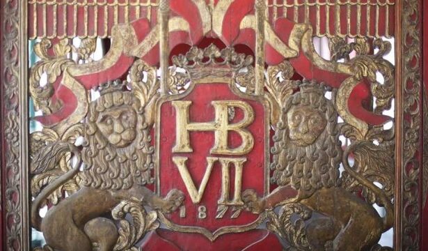 Ornate gold heraldic panel with a crown, the monogram 'HB VII', and two lion supporters on a red background.