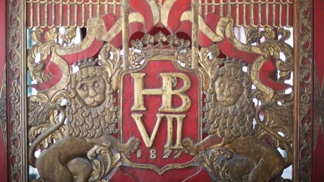 Ornate gold heraldic panel with a crown, the monogram 'HB VII', and two lion supporters on a red background.